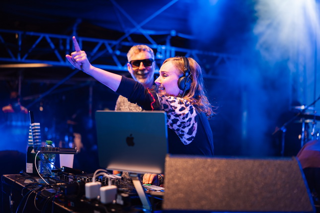 Eine DJ-Performance auf einer Bühne mit Nebel. Eine Person mit Kopfhörern steht hinter einem DJ-Pult und hebt den Arm. Auf dem Pult steht ein Laptop und Wasserflaschen sind sichtbar.