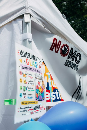 Ein Stand mit einem weißen Zelt. An einer Seite hängt ein farbiger Flyer mit der Aufschrift 'KOMPLIMENTE-GLAS. Nimm eins, Gib eins.' Daneben sind bunte Luftballons in Blau. Eine Logo mit 'NOMO' ist sichtbar.