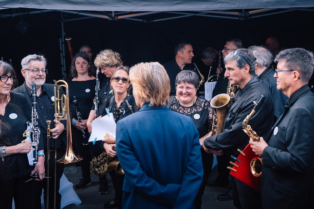 Eine Gruppe von Musikern steht unter einem Zelt. Die Musiker tragen schwarze Kleidung und halten verschiedene Instrumente, darunter Trompeten und Saxophone. Eine Person mit blonden Haaren steht mit dem Rücken zur Kamera und spricht mit der Gruppe, während einige Musiker Notenblätter in den Händen halten. Der Hintergrund ist dunkel, was den Fokus auf die Gruppe lenkt.
