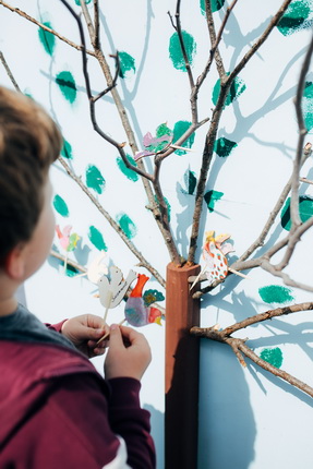 Ein Kind steht vor einem bemalten Baum aus Ästen an einer Wand. Es befestigt farbige, papierene Vögel und Blätter an den Ästen. Die Wand ist hellblau und mit grünen Blättern bemalt.