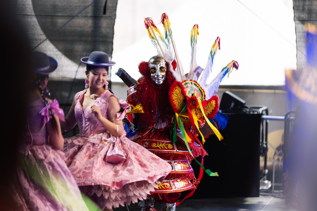 Eine bunte Tanzaufführung, bei der zwei Personen in traditionellen Kostümen auf einer Bühne stehen. Die linke Person trägt ein rosa Kleid und einen Hut, während die rechte Person ein aufwendiges, farbenfrohes Kostüm mit Federn und Verzierungen trägt. Der Hintergrund zeigt eine dunkle Bühne mit einem verschwommenen technischen Bereich.