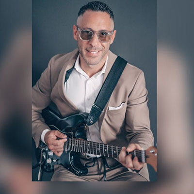 Ein Mann sitzt und spielt eine schwarze Gitarre. Er trägt einen beigen Anzug mit einem weißen Hemd und hat eine Krawatte. Sein Gesicht ist nicht sichtbar.