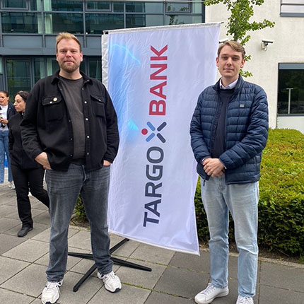 Zwei Männer stehen nebeneinander vor einem Banner der TARGOBANK. Der Mann links trägt ein schwarzes Hemd und Jeans, der Mann rechts einen blauen Parka und helle Hosen. Im Hintergrund sind einige Personen und Gebäude sichtbar.