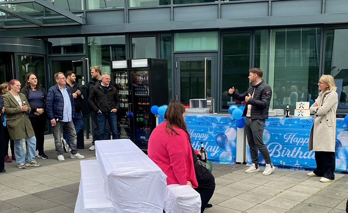 Eine Gruppe von Menschen steht vor einem Tisch, der mit blauen Ballons und einem Banner mit der Aufschrift "Happy Birthday" dekoriert ist. Eine Person spricht, während die anderen zuhören. Im Hintergrund sind Getränkeautomaten sichtbar. Es ist eine Feier im Freien.