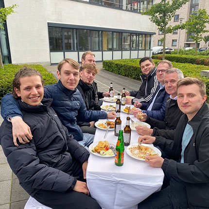 Eine Gruppe von Personen sitzt an einem Tisch im Freien und genießt eine Mahlzeit. Sie haben verschiedene Gerichte auf ihren Tellern und Getränke vor sich. Die Umgebung ist grün mit Bäumen und modernen Gebäuden im Hintergrund. Einige Personen tragen sportliche Kleidung, während andere lässige Outfits tragen. Das Wetter scheint mild zu sein.