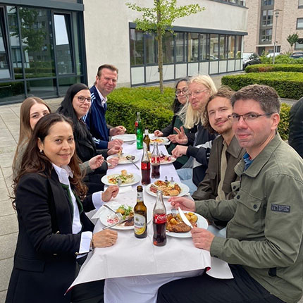 Eine Gruppe von Personen sitzt an einem Tisch im Freien, umgeben von Büschen. Sie haben Teller mit Essen und Getränke wie Bier und Cola. Einige Personen posieren mit den Händen. Die Atmosphäre wirkt gesellig.