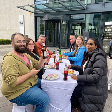 Eine Gruppe von fünf Personen sitzt an einem Tisch im Freien, der mit einem weißen Tuch bedeckt ist. Einige Personen halten Flaschen und Gläser. Auf dem Tisch sind verschiedene Getränke und Snacks sichtbar. Im Hintergrund befindet sich ein modernes Gebäude mit einer Glastür.