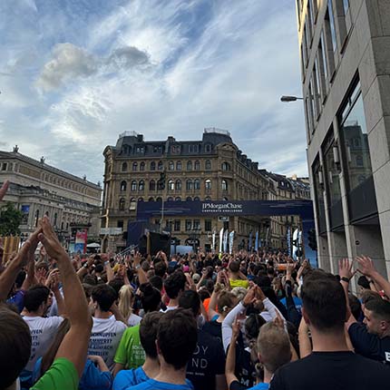 Eine große Menschenmenge steht auf einer Straße, die Hände in die Luft gehoben. Im Hintergrund ist ein historisches Gebäude zu sehen, und ein Banner mit "JPMorgan Chase" hängt über der Versammlung. Der Himmel ist teilweise bewölkt.