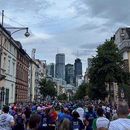Eine große Gruppe von Läufern bewegt sich eine Straße entlang, mit hohen Gebäuden im Hintergrund. Die Teilnehmer tragen bunte Kleidung, einige mit Aufschriften. Der Himmel ist bewölkt, und es gibt Bäume entlang der Straße.