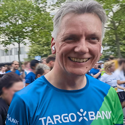 Ein Mann steht im Vordergrund, trägt ein blau-grünes T-Shirt mit dem Logo der TARGOBANK. Im Hintergrund sind andere Läufer und eine grüne Umgebung zu sehen.