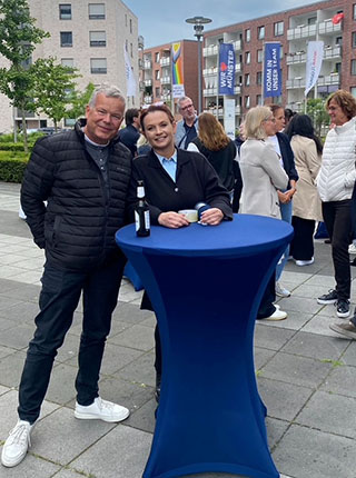 Zwei Personen stehen an einem blauen Stehtisch im Freien. Eine Person trägt eine schwarze Jacke und weiße Sneakers, während die andere einen dunklen Anzug trägt. Im Hintergrund sind weitere Personen und eine Stadtlandschaft mit Flaggen zu sehen.