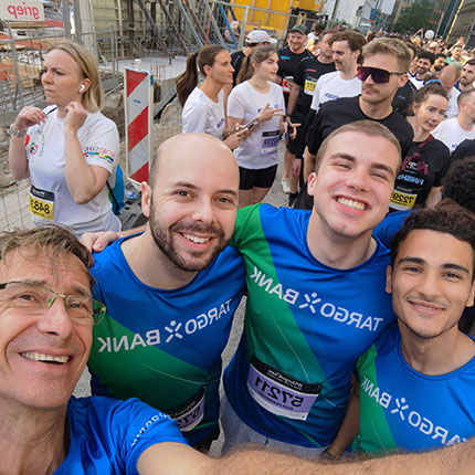 Eine Gruppe von Menschen steht an einem Veranstaltungsort, möglicherweise für ein Rennen. Einige Personen tragen farbige T-Shirts mit dem Logo "TARGOBANK". Im Vordergrund sind mehrere Gesichter sichtbar, während der Hintergrund mit weiteren Teilnehmern gefüllt ist. Die Umgebung sieht nach einer städtischen Baustelle aus.