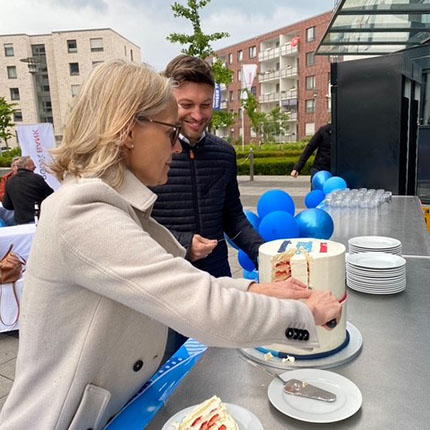 Eine Person mit blonden Haaren schneidet einen mehrstöckigen Kuchen an einem Tisch im Freien. Im Hintergrund sind moderne Gebäude und blaue Luftballons sichtbar. Eine weitere Person steht daneben und beobachtet.