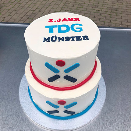 Eine zweistöckige Torte mit einer weißen Zuckerglasur. Auf der Oberseite steht in blauer und schwarzer Schrift "1. JAHRT TDG MÜNSTER". Die untere Torte hat eine rote und eine blaue Dekoration, die symmetrisch angeordnet ist.