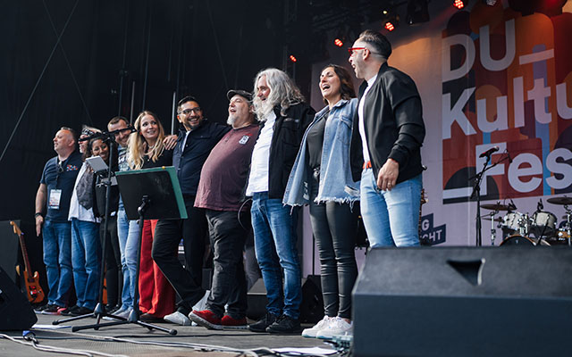 Eine Gruppe von Menschen steht auf einer Bühne beim Du Kultur Festival. Sie stehen zusammen, einige tragen Jeans und T-Shirts, während andere formellere Kleidung tragen. Im Hintergrund sind Scheinwerfer und eine große Banner mit dem Schriftzug 'Du Kultur Festival' zu sehen. Es gibt auch ein Mikrofonständer auf der Bühne.