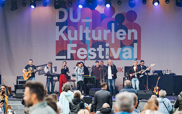 Eine Band tritt auf einer Bühne beim Kulturfestival auf. Im Hintergrund ist ein großes Banner mit der Aufschrift "DU Kulturlen Festival" zu sehen. Die Musiker spielen Instrumente wie Gitarre und Keyboard, während die Menge vor der Bühne zuschaut. Es gibt eine vielfältige Gruppe von Menschen im Publikum, die sich für die Darbietung interessieren.