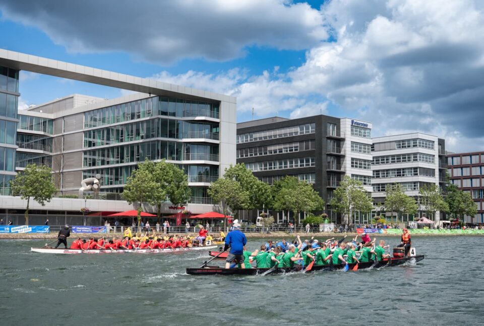 Zwei Drachenboot-Teams paddeln auf einem Gewässer vor modernen Gebäuden. Eine Gruppe in roten Shirts und eine andere in grünen Shirts. Im Hintergrund sind Bäume und Terrassen mit roten Sonnenschirmen zu sehen. Der Himmel ist teils bewölkt.