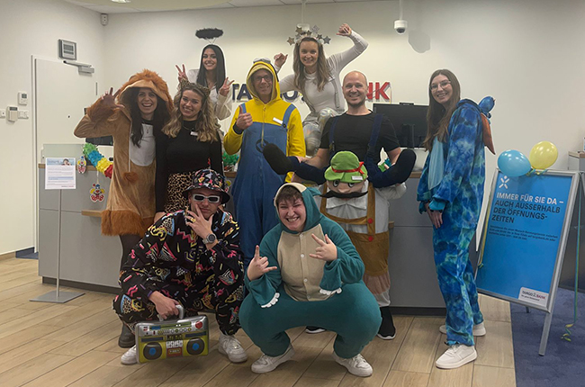 Eine Gruppe von zehn Personen in bunten Kostümen steht im Empfangsbereich. Einige tragen Tierkostüme, andere haben bunte Overalls an. Eine Person hält ein großes Kassettenradio. Im Hintergrund ist ein Empfangstresen mit einem Schild und Luftballons zu sehen.