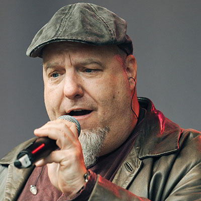 Ein Mann mit einer grauen Mütze und einem Lederjacke hält ein Mikrofon. Sein Gesicht ist unscharf, aber er scheint auf einer Bühne zu singen.