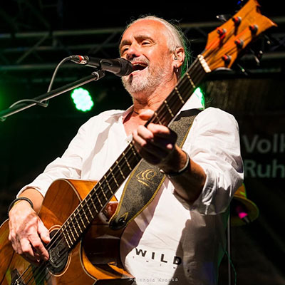 Ein Musiker steht auf einer Bühne und spielt Gitarre. Er trägt ein weißes Hemd und hat seine Gitarre in der Hand. Im Hintergrund sind Lichter zu sehen.