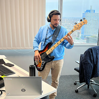 Ein Musiker steht in einem modernen Büro und spielt Bassgitarre. Er trägt Kopfhörer und ein hellblaues Sweatshirt. Vor ihm steht ein Laptop und eine Thermoskanne, während im Hintergrund eine große Fensterfront mit Stadtansicht zu sehen ist.