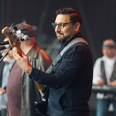 Ein Musiker spielt Bass auf einer Bühne. Er trägt ein schwarzes Oberteil und hat eine Brille auf. Im Hintergrund sind weitere Musiker zu sehen, die Mikrofone benutzen und Instrumente spielen.