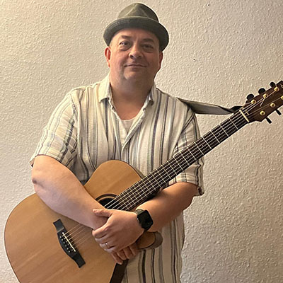 Eine Person steht mit einer Akustikgitarre in der Hand. Die Person trägt ein gestreiftes Hemd und einen grauen Hut. Die Wand im Hintergrund ist hell und strukturiert.