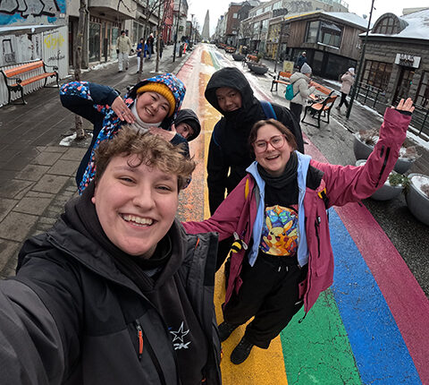 Eine Gruppe von vier Personen posiert in der Mitte einer bunten Straße, die von Graffiti umgeben ist. Die Straße hat eine Regenbogenfarbgestaltung. Im Hintergrund sind weitere Menschen und Geschäfte zu sehen. Die Gruppe macht eine Selfie-Pose, während sie in die Kamera lächelt.