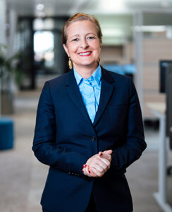 Eine Frau in einem dunkelblauen Blazer und einer hellblauen Bluse steht in einem modernen Büro. Sie lächelt, hat kurze, blonde Haare und trägt goldene Ohrringe. Der Hintergrund zeigt Büroeinrichtungen und einen hellen, offenen Raum.