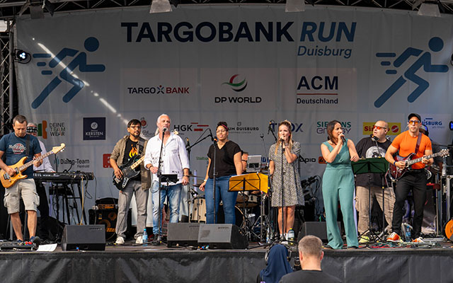 Eine Band steht auf einer Bühne mit einem Banner, das "TARGOBANK RUN Duisburg" zeigt. Sie spielen verschiedene Instrumente, darunter Gitarre und Gesang. Im Hintergrund sind mehrere Logos sichtbar, darunter TARGOBANK und DP WORLD. Die Bandmitglieder tragen unterschiedliche Kleidung und scheinen miteinander zu interagieren.