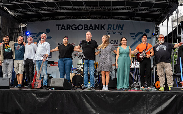 Eine Gruppe von zehn Personen steht auf einer Bühne, die mit Musikinstrumenten und einem Banner für die TARGOBANK RUN Veranstaltung in Duisburg dekoriert ist. Die Menschen stehen nebeneinander und halten sich an den Händen. Einige tragen lässige Kleidung und es gibt eine Gitarre sowie verschiedene Musikinstrumente im Hintergrund. Das Banner im Hintergrund zeigt den Schriftzug 'TARGOBANK RUN'.