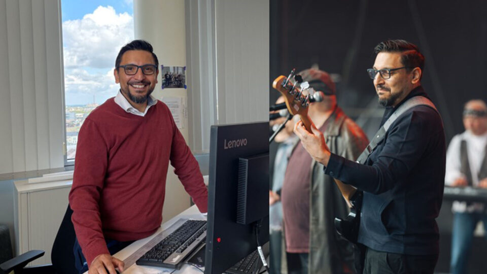 Ein Mann in einem roten Pullover steht an einem Schreibtisch mit einem Lenovo-Computer. Im Hintergrund sieht man ein Fenster mit einer Aussicht auf eine Stadt. Rechts ist ein anderer Mann auf einer Bühne zu sehen, der ein Bassinstrument spielt, während eine Band im Hintergrund auftritt.
