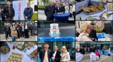 Eine Veranstaltung im Freien mit mehreren Gruppen von Menschen, die auf einem Platz stehen. Im Vordergrund stehen zwei Personen vor einem Banner der Ergo Bank. In der Mitte eine Gruppe von Personen, die lächeln und posieren. Eine festliche Torte mit dem Text „Happy Birthday“ und einer Dekoration in Blau und Rot. Es gibt verschiedene Speisen auf einem Tisch, darunter Brote, Brötchen und Salate, sowie Getränkestationen. Im Hintergrund sind Gebäude und Bäume sichtbar.