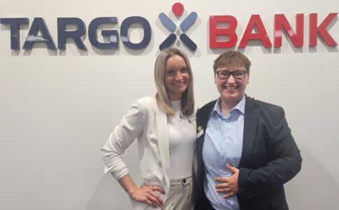 Zwei Personen stehen vor einer Wand mit dem Logo der TARGOBANK. Die Frau links trägt einen weißen Blazer und hat lange blonde Haare. Die Person rechts trägt einen dunklen Anzug und ein hellblaues Hemd. Beide Personen lächeln.