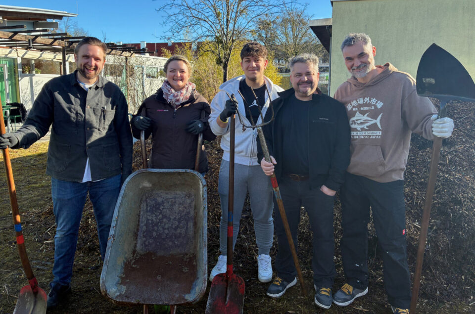 Fünf Personen stehen in einem Garten. Sie halten Schaufeln und eine Schubkarre. Im Hintergrund ist ein Haufen aus Holzschnitzeln und Bäume. Die Sonne scheint.