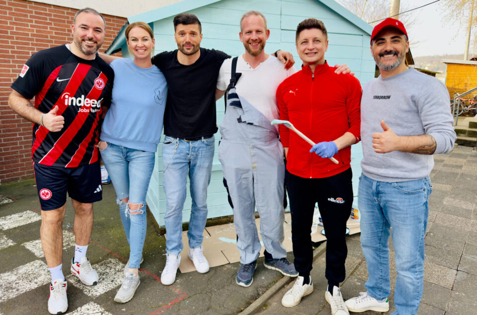 Sechs Personen stehen vor einem blauen Holzschuppen. Die Männer tragen Sportkleidung, einer ist in einem rot-schwarzen Trikot, ein anderer in einem roten Sweatshirt. Eine Frau trägt ein hellblaues Oberteil und Jeans. Alle lächeln und posieren mit Daumen hoch. Im Hintergrund sind einige Werkzeuge und Malutensilien sichtbar.