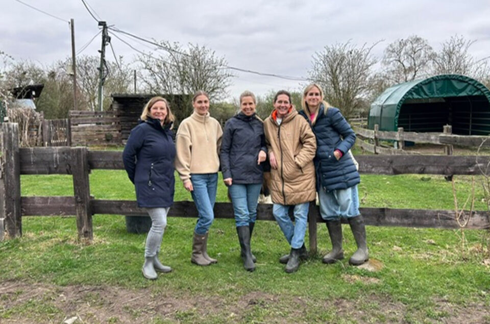 Fünf Frauen stehen an einem Holzzaun auf einer Wiese. Sie tragen warme Kleidung und Gummistiefel. Im Hintergrund sind Bäume und eine grüne Plane zu sehen.