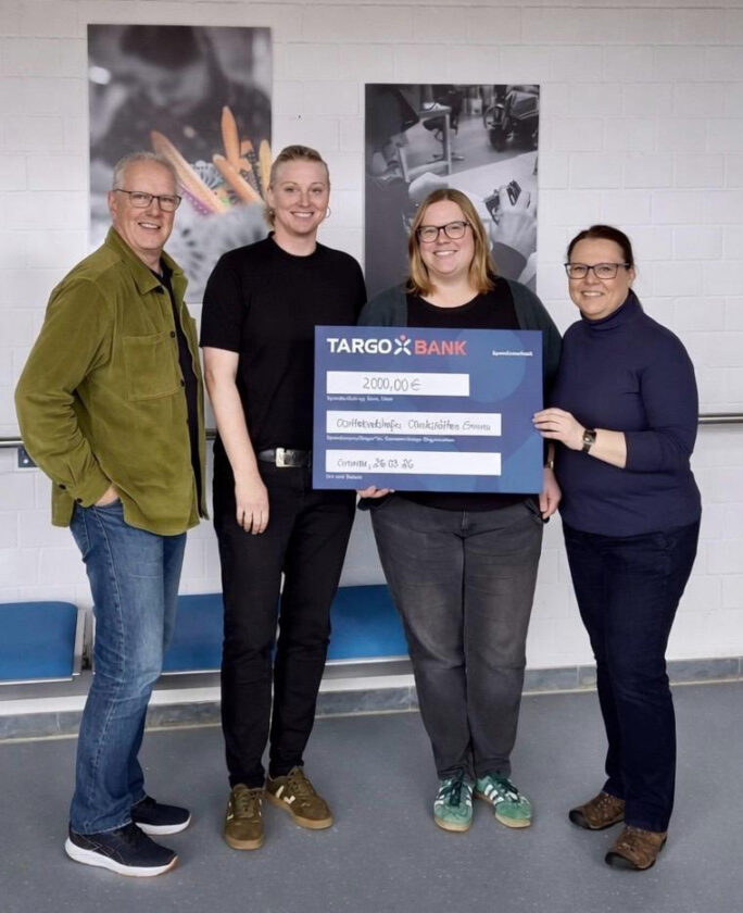 Vier Personen stehen zusammen in einem Raum, halten einen großen Scheck von TARGOBANK über 2.000,00 €. Im Hintergrund sind kreative Materialien sichtbar.