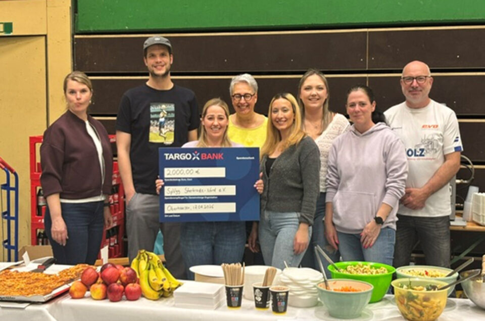 Eine Gruppe von sieben Personen steht vor einem Tisch mit Essen und Getränken. Sie halten einen großen Scheck der TARGOBANK über 2000,00 €, der an den Verein "Spiels Sternenkinder e.V." geht. Auf dem Tisch sind Obst, Salate und Snacks angerichtet.