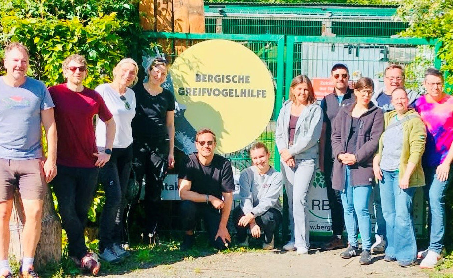 Eine Gruppe von Menschen steht vor einem Zaun mit einem gelben Schild, auf dem 'Bergische Greifvogelhilfe' steht. Es sind insgesamt 13 Personen zu sehen, die lächeln und in lockerer Kleidung gekleidet sind. Der Hintergrund zeigt eine grüne Umgebung mit Pflanzen und Bäumen.