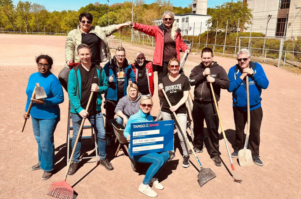 Eine Gruppe von 12 Personen steht auf einem Sportplatz. Sie halten verschiedene Werkzeuge wie Schaufeln und Besen. Eine Person sitzt in einem Schubkarren und hält ein großes Scheck-Schild der TARGOBANK über 2.000 Euro, das an den FC Pohl-Lap-Wess wird. Die Szene ist sonnig und freundlich.