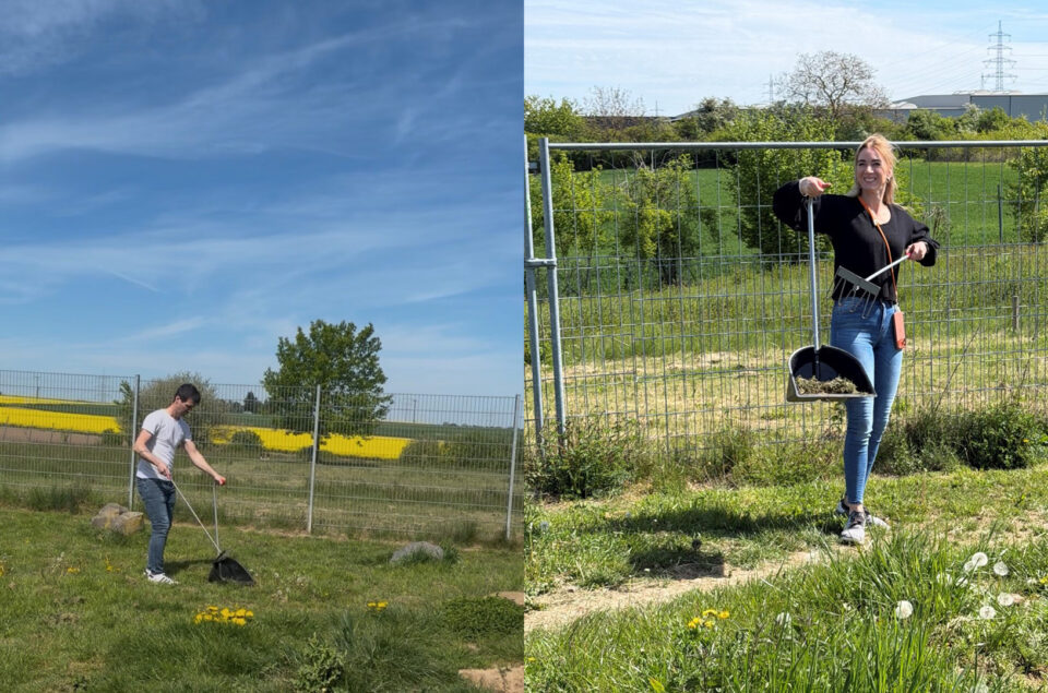 Ein Mann steht auf der linken Seite des Bildes, während er mit einer Schaufel in einem grünen Feld arbeitet. Auf der rechten Seite steht eine Frau, die mit einer Schaufel und einer Harke in der Hand lächelt. Beide stehen hinter einem Drahtzaun, umgeben von Gras und wenigen Blumen. Im Hintergrund sind Bäume und ein blauer Himmel mit einigen Wolken sichtbar.