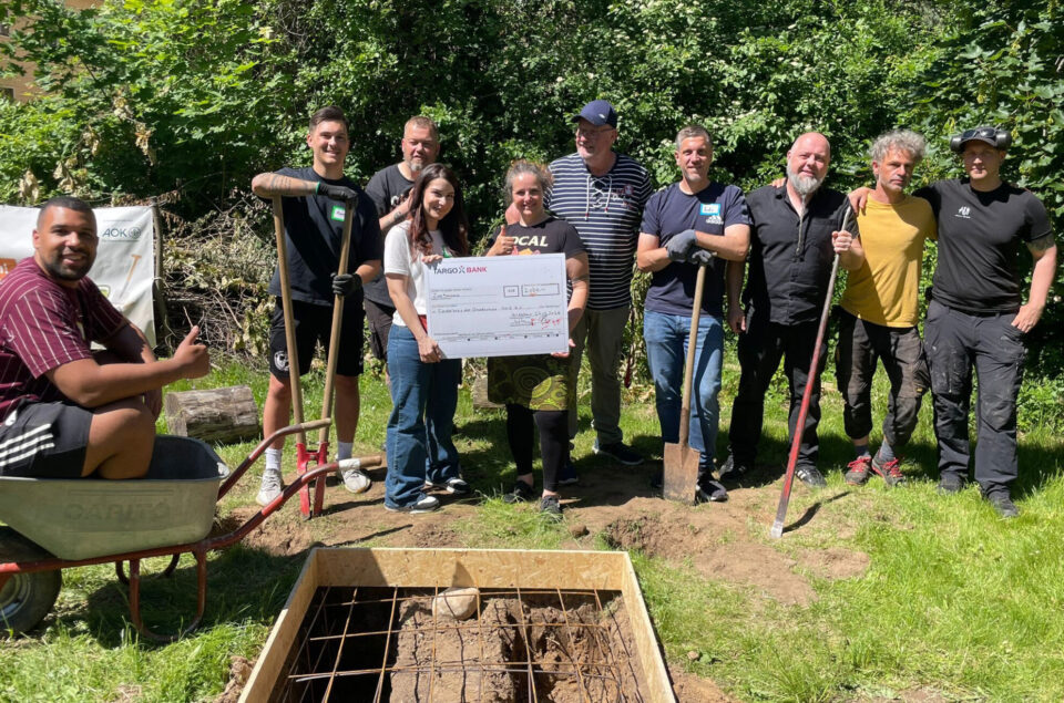 Eine Gruppe von neun Personen steht in einem Garten, einige mit Schaufeln und Werkzeugen. Eine Person hält einen großen Scheck von der 'ARGO BANK'. Im Vordergrund ist eine Holzbox mit einem Gitter zu sehen.