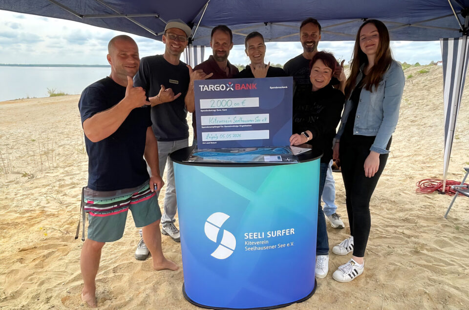 Eine Gruppe von sechs Personen steht an einem Sandstrand unter einem Zelt. Sie halten einen großen Scheck von 2.000 Euro für den Kiteverein Seelhausen See e.V. in der Hand. Die Personen lächeln und zeigen Daumen hoch. Im Hintergrund ist der See sichtbar, und der Himmel ist bewölkt.