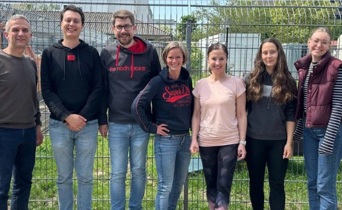 Eine Gruppe von sieben Personen steht vor einem Zaun. Von links nach rechts: ein Mann in einem dunklen Oberteil, ein Mann in einem schwarzen Hoodie mit dem Logo "the north face", ein Mann mit Brille, eine Frau mit kurzem Haar in einem schwarzen Oberteil, eine Frau in einem hellen T-Shirt, eine Frau mit langen, lockigen Haaren in einem schwarzem Oberteil, und eine Frau mit Brille in einer dunkelroten Weste. Der Hintergrund zeigt eine grüne Wiese und einige Gebäude.