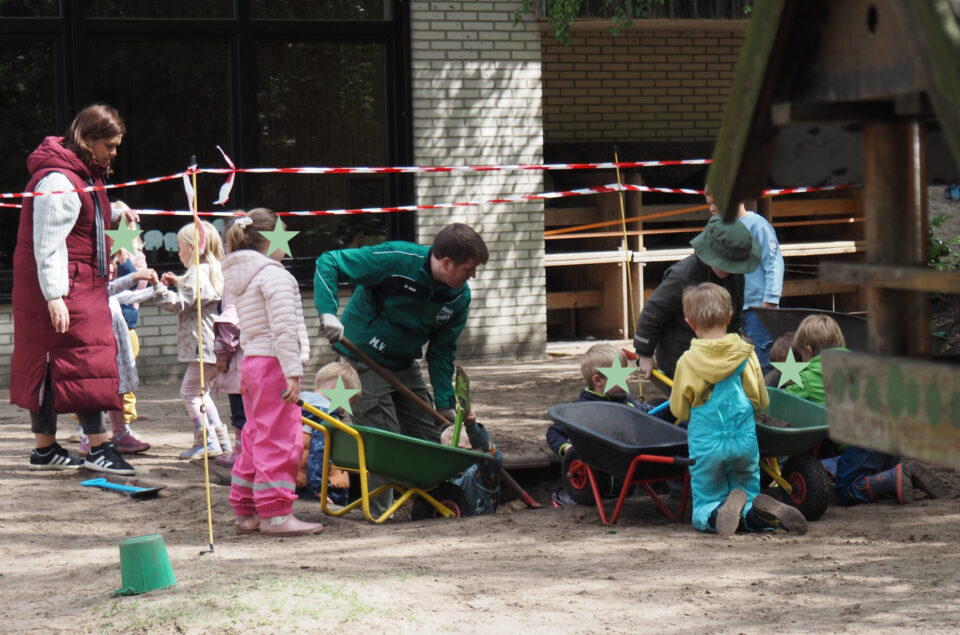 Eine Gruppe von Kindern spielt im Freien, während ein Erwachsener hilft. Einige Kinder sitzen in Schubkarren, andere graben im Sand. Ein rotes und weißes Absperrband ist sichtbar. Die Kinder tragen bunte Kleidung, während der Erwachsene einen grünen Arbeitsanzug hat.