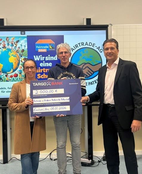 Drei Personen stehen vor einem Bildschirm, der Informationen über Fairtrade zeigt. Eine Frau links trägt eine Brille und einen langen schwarzen Mantel. Der Mann in der Mitte hält einen großen Scheck von 2.000 Euro, der an die 'Freunde und Partner der Realschule' gerichtet ist. Der Mann rechts trägt einen schwarzen Anzug und lächelt. Das Datum auf dem Scheck ist der 08.05.2026.