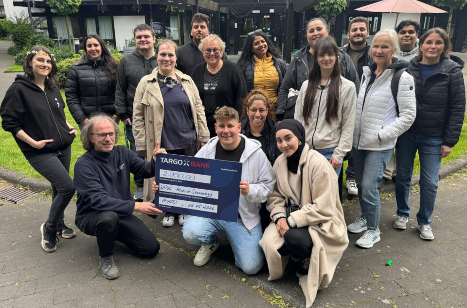 Eine Gruppe von Menschen posiert im Freien. In der Mitte hält eine Person einen großen Scheck der TARGOBANK über 2.000,00 Euro. Die Gruppe besteht aus Männern und Frauen unterschiedlichen Alters, die freundlich in die Kamera lächeln. Im Hintergrund sind Bäume und ein überdachter Bereich sichtbar.