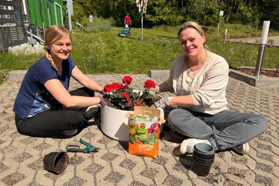 Zwei Frauen sitzen auf dem Boden und pflanzen rote Blumen in einen weißen Blumentopf. Eine Frau trägt ein blaues T-Shirt und hat eine Zopffrisur, während die andere ein helles Sweatshirt anhat. Neben ihnen liegt eine offene Tüte Blumenerde und mehrere schwarze Töpfe. Im Hintergrund sind grüne Wiesen und eine Spielplatzstruktur sichtbar.