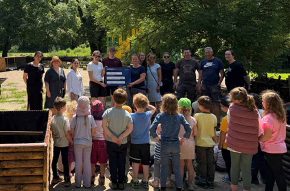 Eine Gruppe von Erwachsenen und Kindern steht im Freien. Die Erwachsenen halten ein großes Schild mit Aufschriften. Im Hintergrund sind Bäume und eine Spielplatzstruktur sichtbar. Die Kinder stehen in mehreren Reihen und schauen zur Gruppe der Erwachsenen.